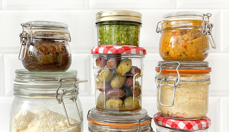 Collection of glass storage jars with various preserves and pickled items, including olives, pesto, and other colourful condiments, some with traditional checkered jar lids