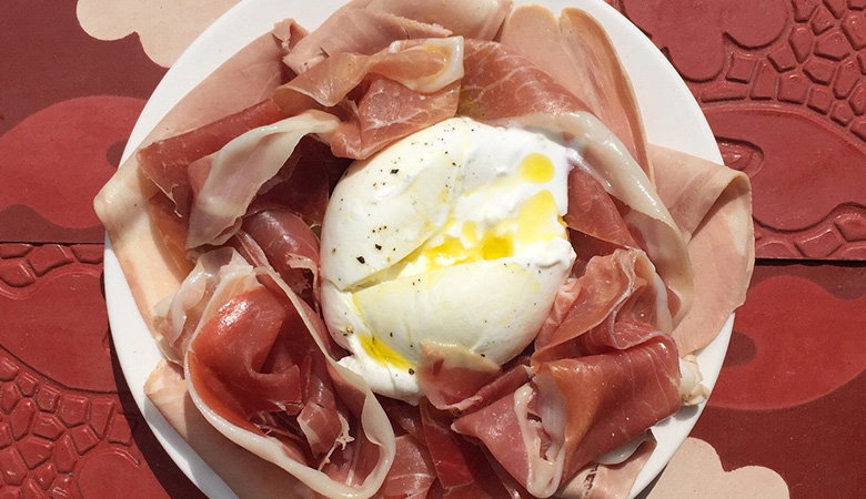 Gourmet appetizer plate with fresh burrata or mozzarella cheese topped with a golden egg yolk, surrounded by delicate slices of prosciutto on a white plate