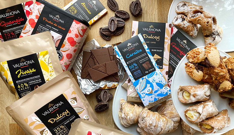 Premium chocolate and confection display featuring Valrhona chocolate bars, artisanal pastries including cannoli, and various luxury sweets arranged on a wooden surface