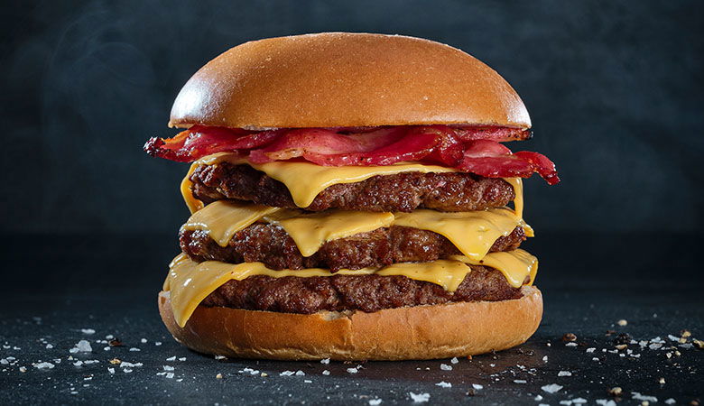Towering triple-patty cheeseburger with melted cheese, crispy bacon, and beef patties on a brioche bun, photographed against a dark slate background