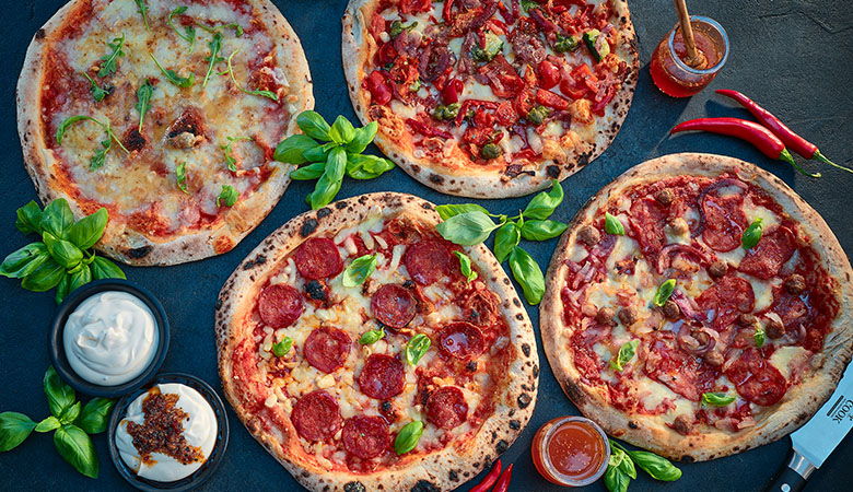Artisanal wood-fired pizzas with various toppings including pepperoni, vegetables, and herbs, accompanied by basil leaves, chilli peppers, and dipping sauces on a dark surface