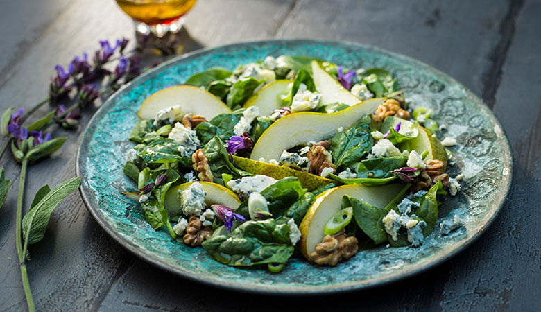 Gourmet salad featuring mixed greens, sliced pears, crumbled blue cheese, walnuts, and edible purple flowers on a rustic teal ceramic plate with olive oil drizzle