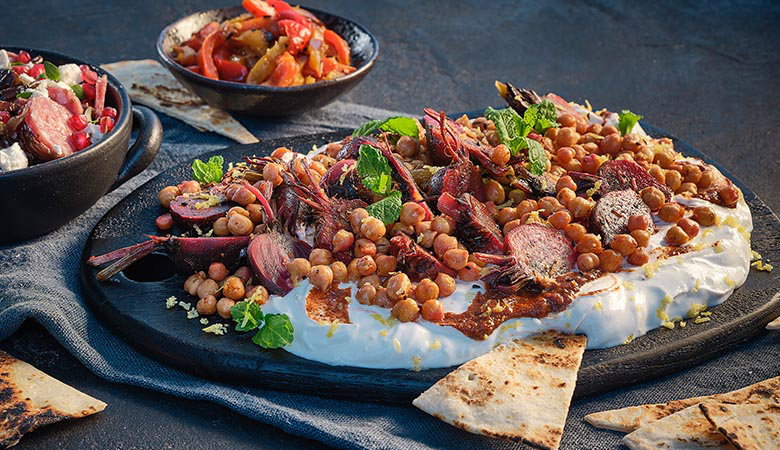 A vibrant platter of roasted chickpeas, colourful vegetables, and greens on creamy yogurt garnished with mint, alongside bowls of salads and pita slices.