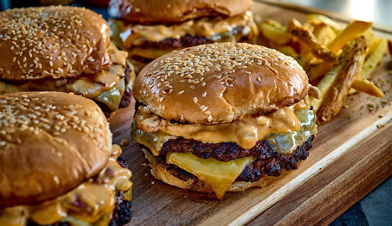 Juicy cheeseburgers with sesame seed buns and creamy sauce on a wooden board, accompanied by crispy golden fries, create a hearty, appetising scene.