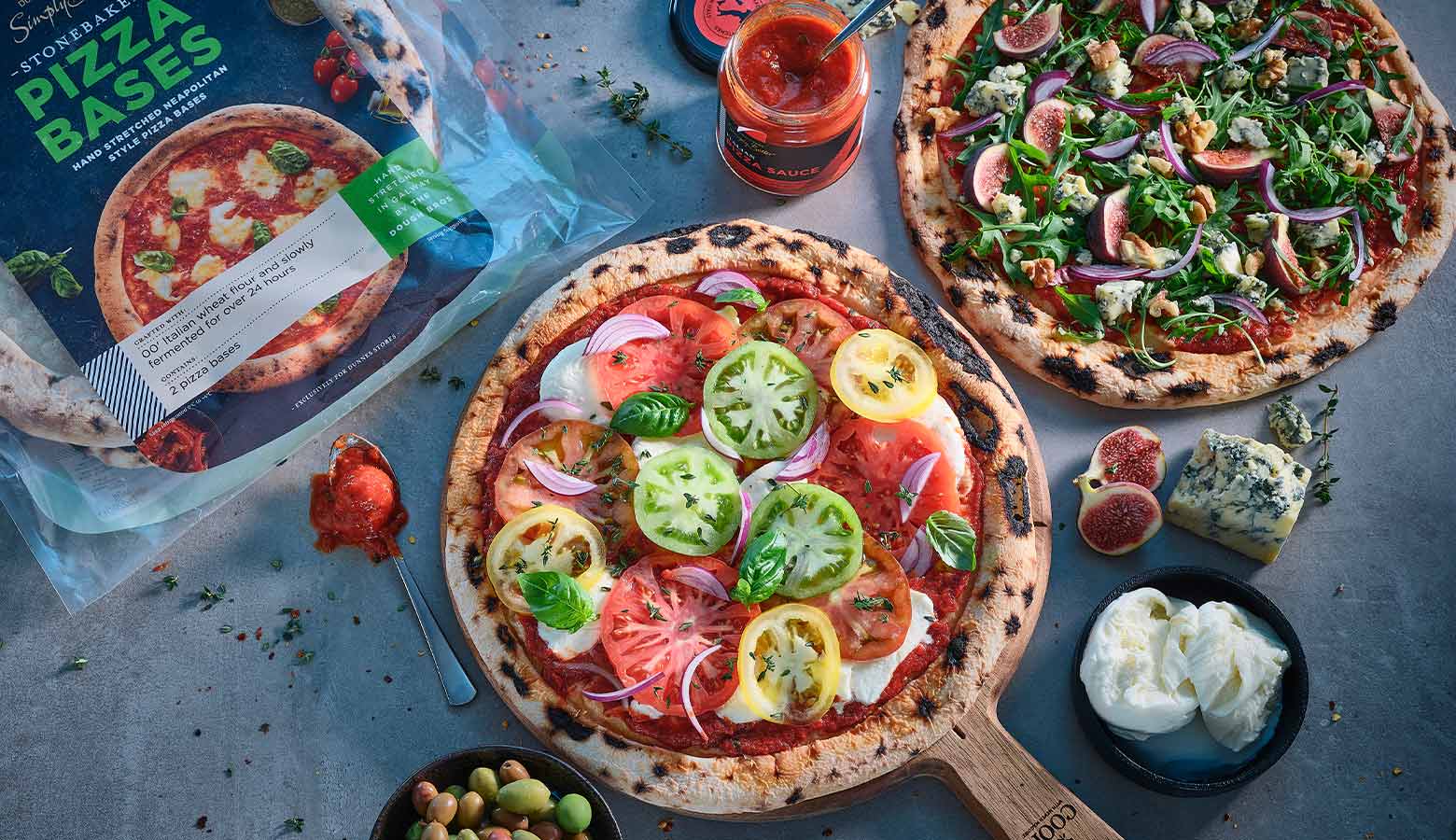 Two fresh pizzas on a table: one with tomatoes and basil, the other with figs and arugula. Ingredients like cheese and sauce surround them. Inviting and colourful.