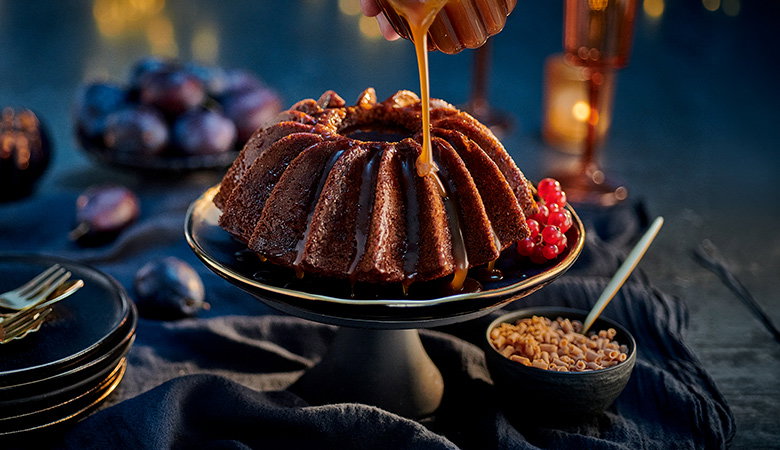 Sticky toffee pudding on a cake stand with warm toffee sauce being poured over the top, garnished with red berries and surrounded by dates in a moody dining setting.