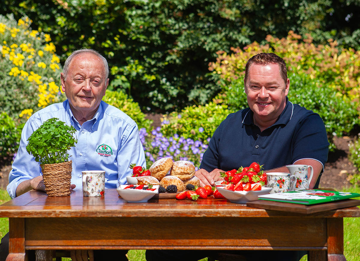 Pat Clarkle and Neven Maguire sit at a wooden table outdoors, surrounded by blooming flowers, enjoying a spread of fresh strawberries, scones, and tea.
