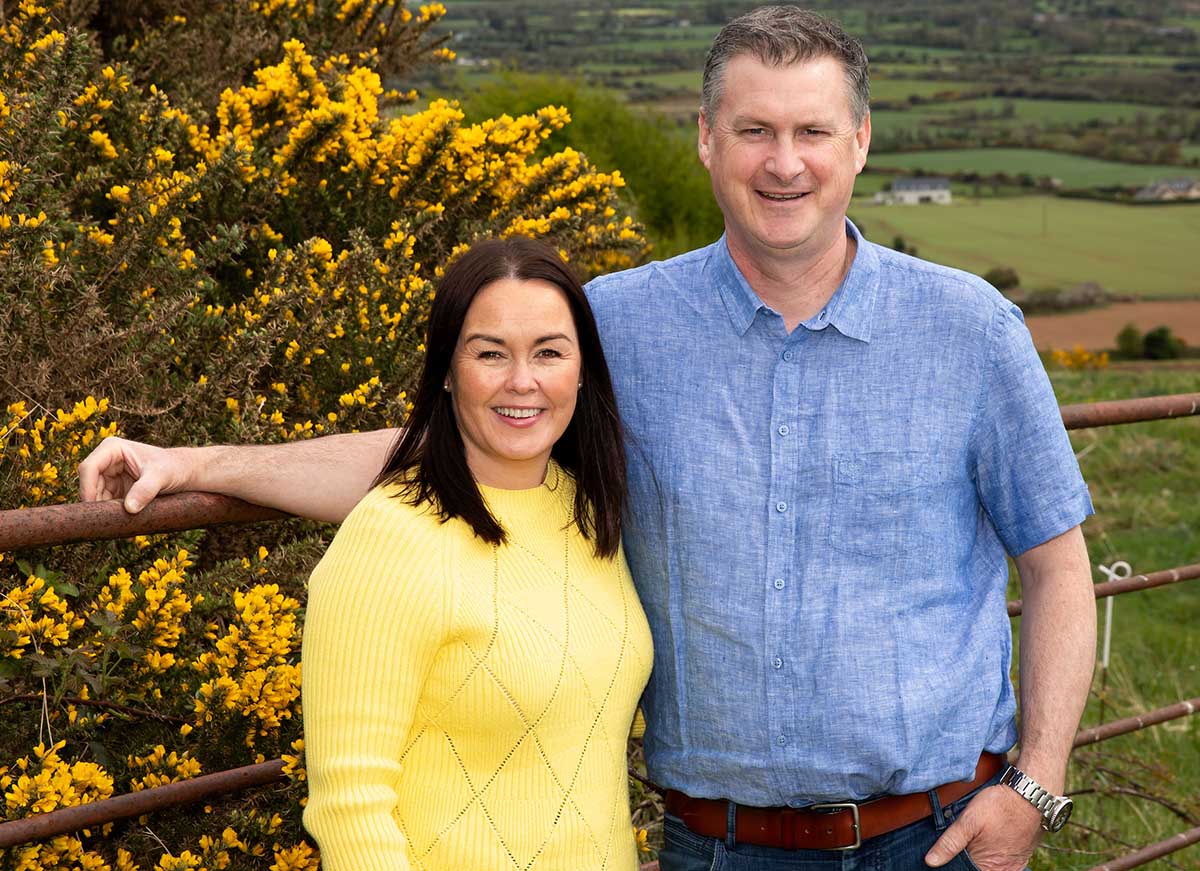 A couple stands together outdoors, surrounded by vibrant yellow flowers and a scenic green landscape. The man is wearing a blue shirt, while the woman is in a yellow sweater.