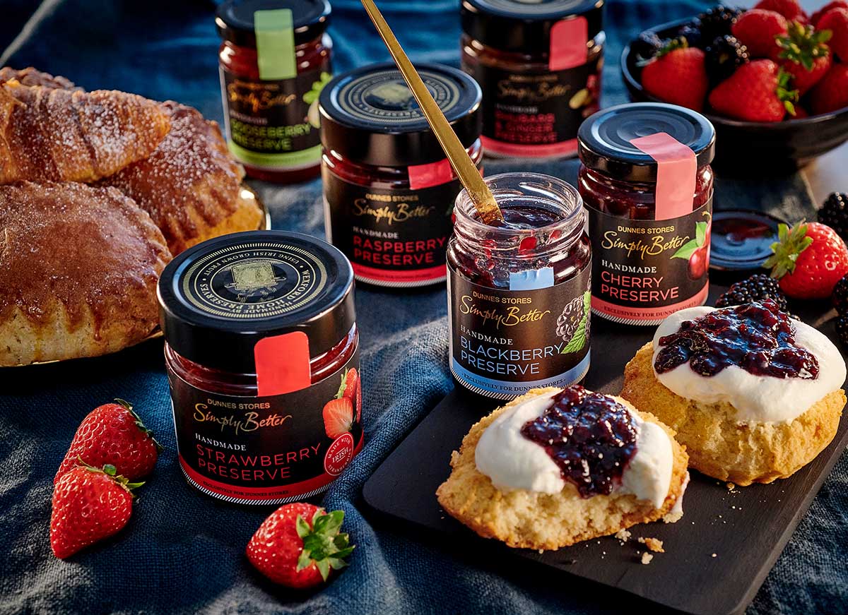 A selection of handmade fruit preserves beside fluffy scones topped with cream and jam, alongside fresh berries and croissants.