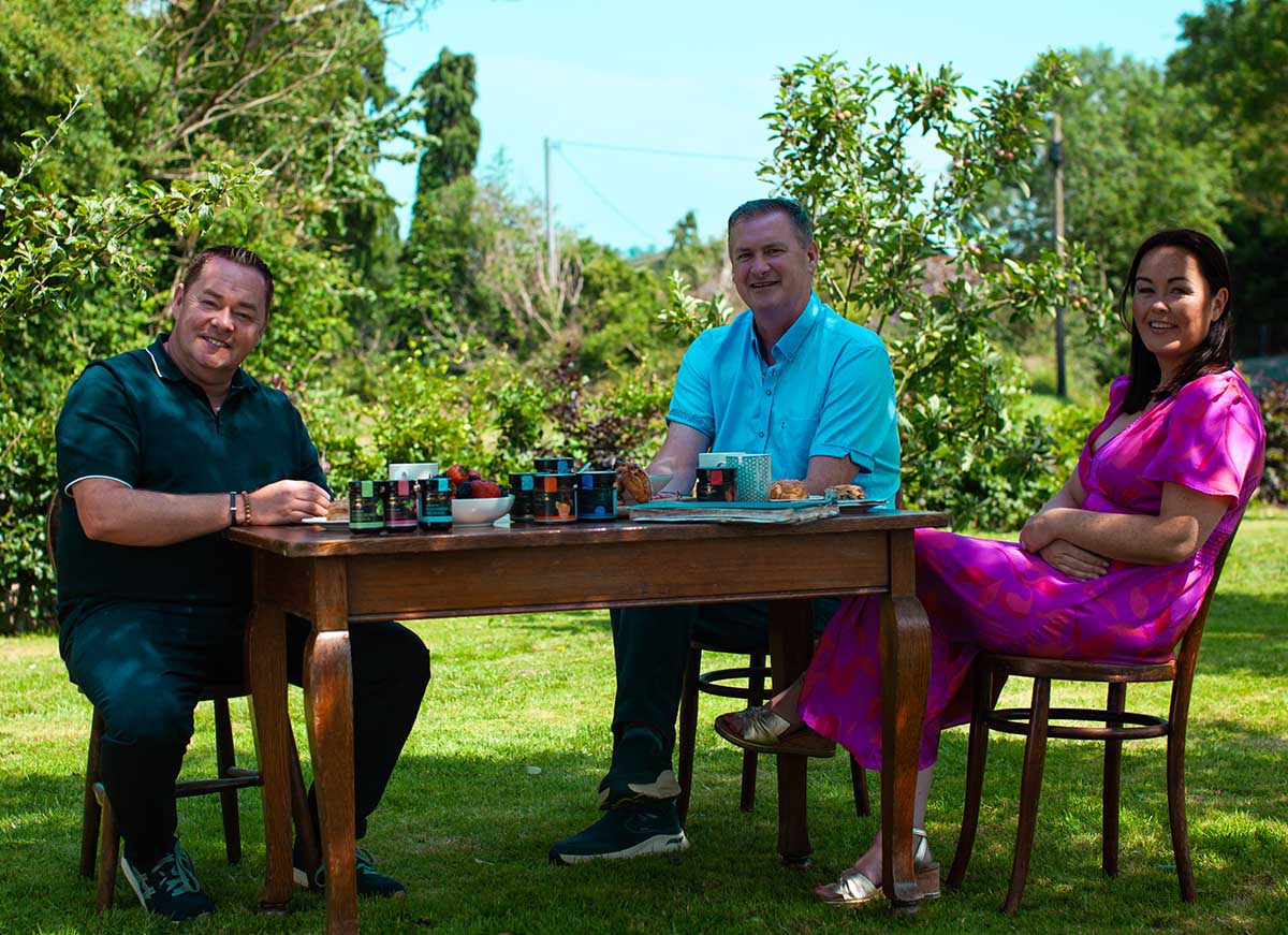 Neven Maguire sits with Tom and Laura Sinnott at a wooden table filled with snacks and colourful containers in a lush garden setting.