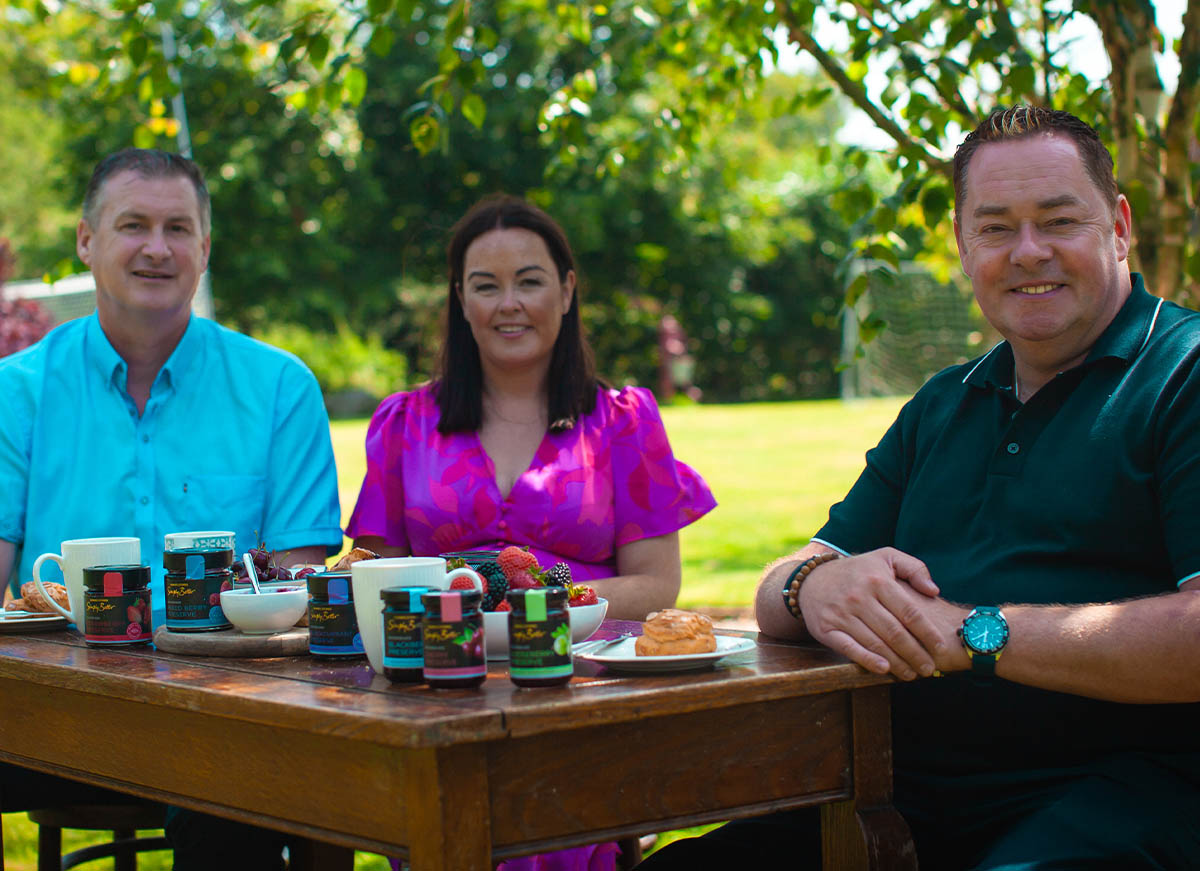 Laura, Tom and Neven Maguire are seated at a wooden table outdoors, surrounded by greenery, with an assortment of jars and food items in front of them.