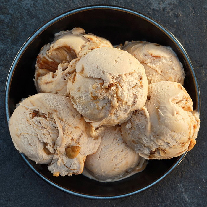 A close-up of a bowl filled with creamy, caramel coloured ice cream scoops, featuring swirls and nut pieces throughout.