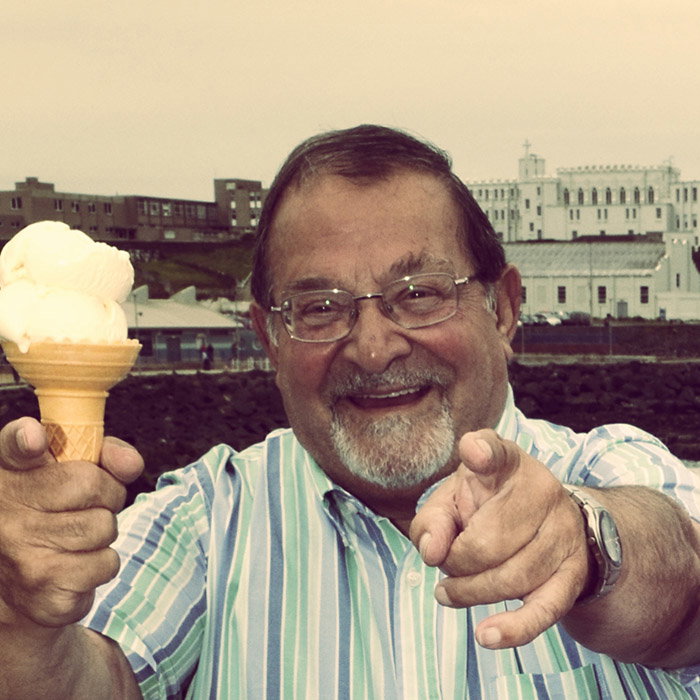 Guido Morelli wearing a striped shirt holds an ice cream cone and points toward the camera, with buildings and rocks in the background
