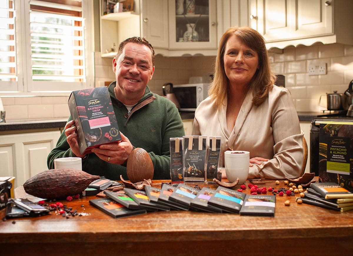 Ann Rudden and Neven Maguire display various chocolate bars and related products on a kitchen table, including a large chocolate egg and cocoa pod.
