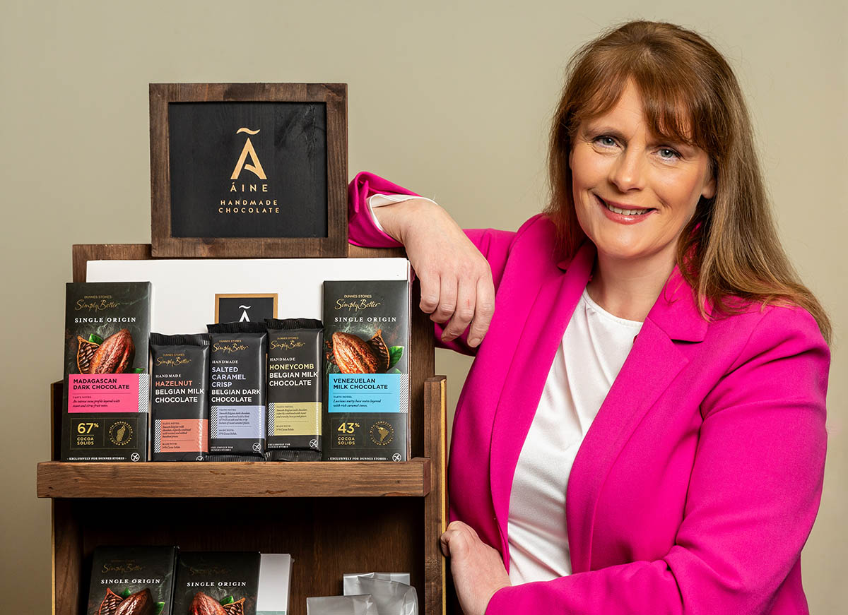 Woman in pink blazer poses beside wooden shelf displaying various dark and milk chocolate bars from &Aacute;ine Handmade Chocolate.