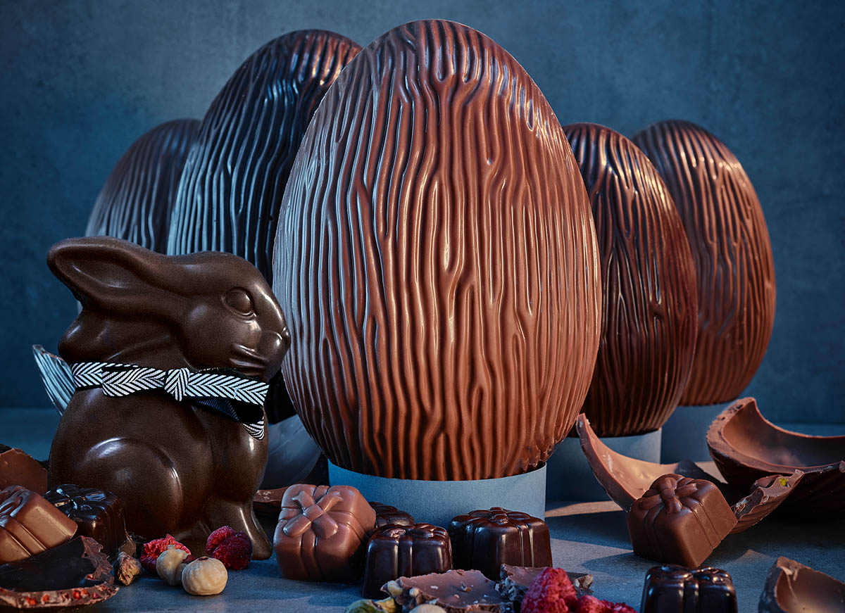 Chocolate Easter eggs and a chocolate bunny with a patterned ribbon, surrounded by assorted chocolates and nuts on a blue surface.