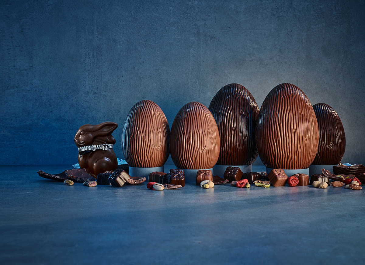 Rows of textured chocolate Easter eggs and a chocolate bunny surrounded by assorted chocolate pieces and nuts on a blue surface.