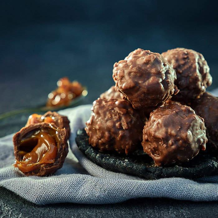 A close-up of chocolate-covered caramel truffles stacked on a black plate, with one truffle cut in half, revealing gooey caramel inside.