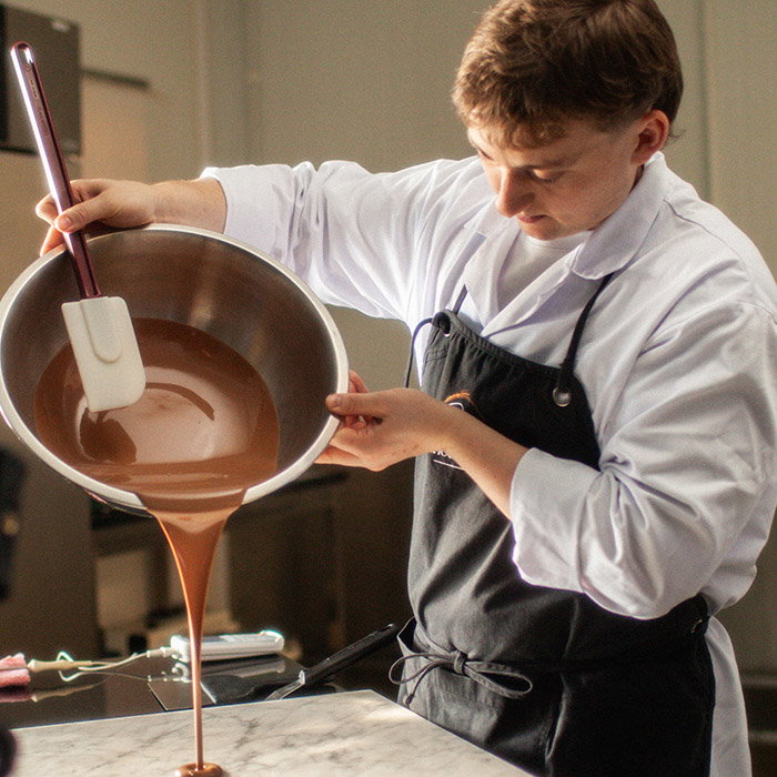A chef in a white coat and black apron pours rich melted chocolate from a bowl, using a spatula, into a marble countertop.