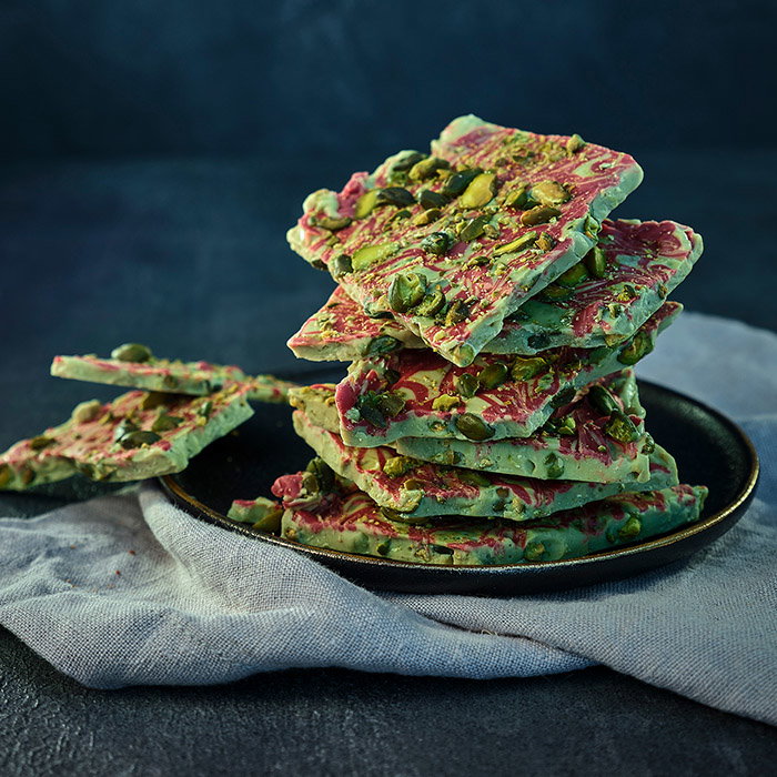 A stack of colourful chocolate bark with swirls of pink and green, topped with crushed pistachios, on a dark textured background.