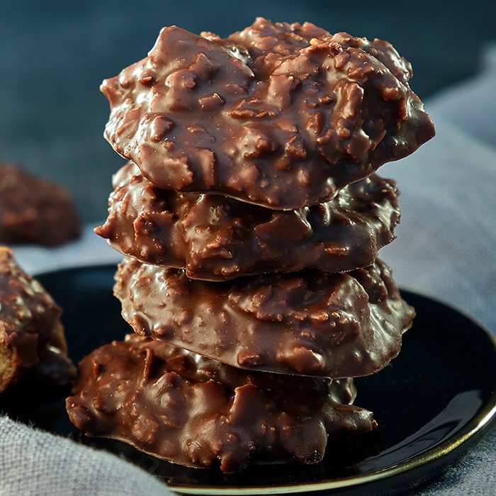 A stack of four chocolate-covered cookies, topped with crispy bits, on a black plate against a soft grey background.
