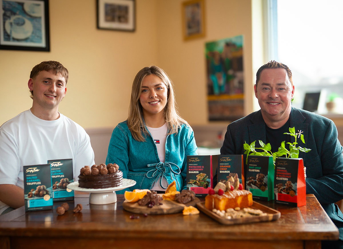 Nevan Maguire visits Bon Chocolatier's HQ and sits at a table, surrounded by various desserts and chocolates, featuring colourful packaging of gourmet treats.