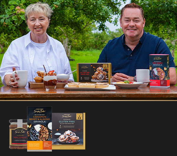 Neven Maguire sits with Siobh&aacute;n from The Foods of Athenry at a wooden table, surrounded by greenery, with various food items and healthy snacks displayed on the table.
