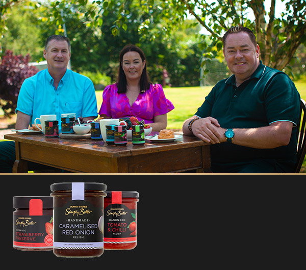 Tom and Laura Sinnott sit with Neven Maguire at a wooden table, showcasing various jars of jams and relishes amid lush greenery.