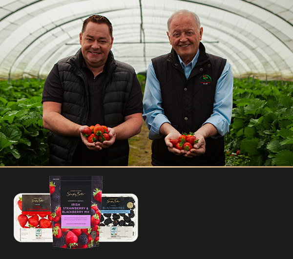 Pat Clarke and Neven Maguire are in a greenhouse holding fresh strawberries while various berry products are displayed in front of them. Lush green plants surround them.