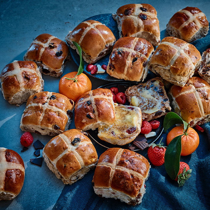 A spread of freshly baked hot cross buns with chocolate, berries, and citrus fruits arranged on a textured surface.