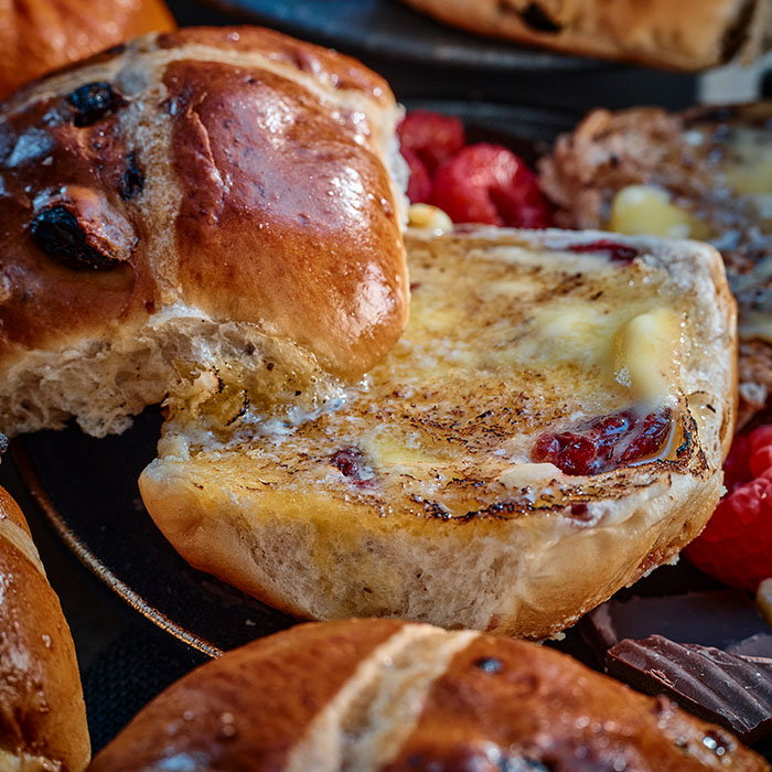 A close-up of a toasted hot cross bun, oozing with butter, surrounded by fresh raspberries and other pastries.