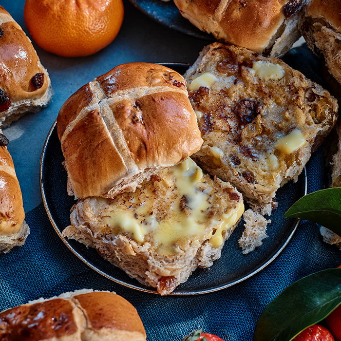 A close-up of hot cross buns, one sliced open and topped with melted butter, surrounded by a tangerine and green leaves.