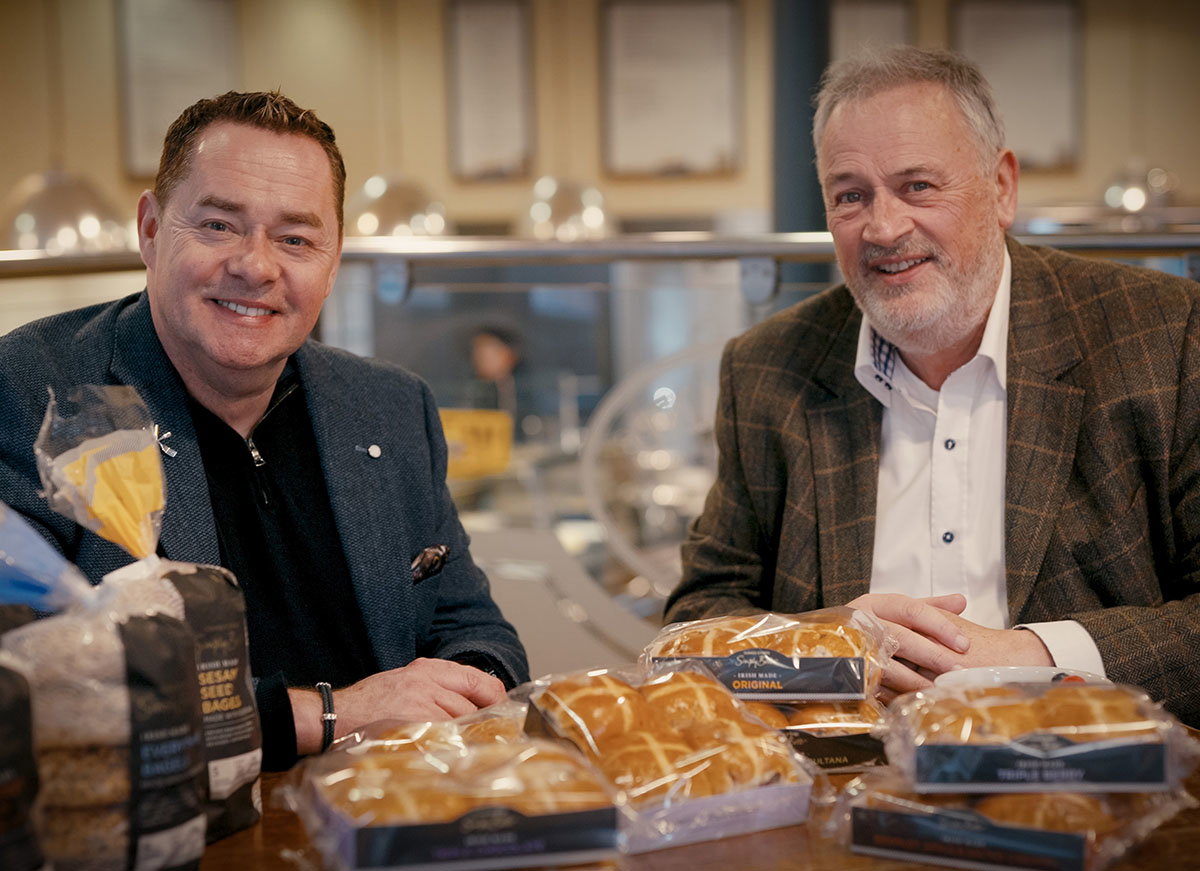 Neven Maguire and Pat McCloskey sit at a table with bags of cross buns, discussing items in front of them in a cozy café setting.