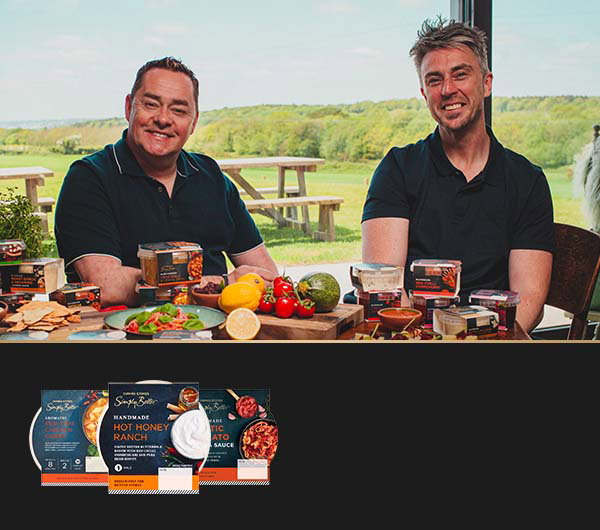 Neven Maguire seated beside Chef Kevin Aherne of Sage at a table displaying packaged food products, fresh vegetables and ingredients, with branded product packaging shown below.