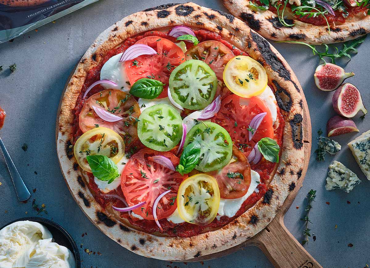 Stone-baked pizza topped with sliced heirloom tomatoes, mozzarella, red onion and fresh basil on a wooden pizza peel.