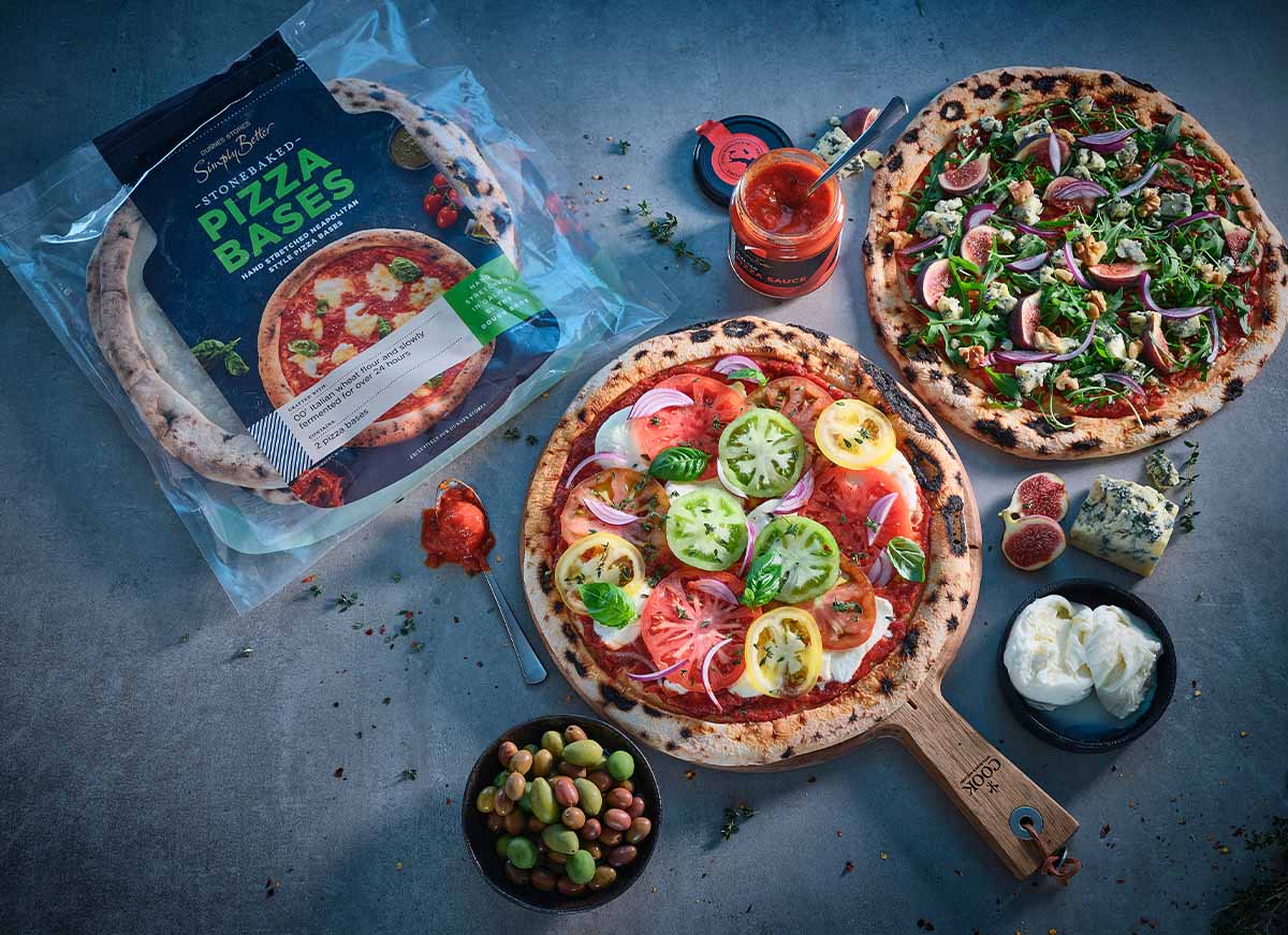Pack of Simply Better Stonebaked Pizza Bases displayed alongside two finished stone-baked pizzas with fresh toppings, olives, mozzarella, blue cheese, figs and tomato sauce.