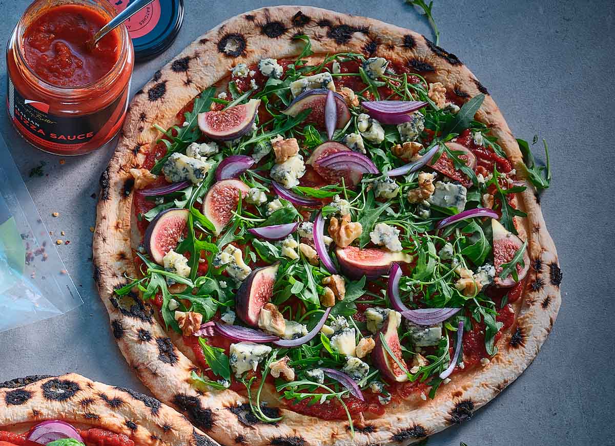 Stone-baked pizza topped with figs, blue cheese, rocket leaves, red onion and walnuts, served beside a jar of pizza sauce.