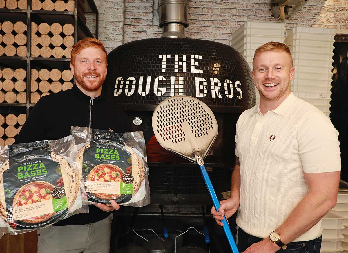 Eugene and Ronan Greaney standing in front of The Dough Bros pizza oven, holding Simply Better Stonebaked Pizza Bases and a pizza peel in a restaurant setting.