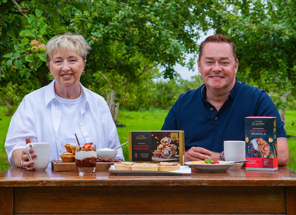 Neven Maguire sits with Siobh&aacute;n from The Foods of Athenry at a wooden table in an outdoor setting displays various desserts and a box of mince pies, surrounded by green trees.