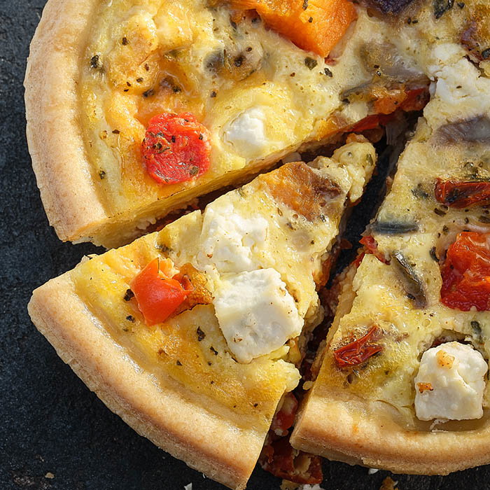 A delicious, partially sliced quiche topped with tomatoes, cheese, and herbs on a golden crust, resting on a dark surface.