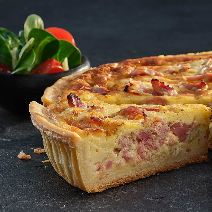 A slice of golden quiche with ham, cheese, and crispy edges, served alongside a fresh salad of greens and cherry tomatoes.