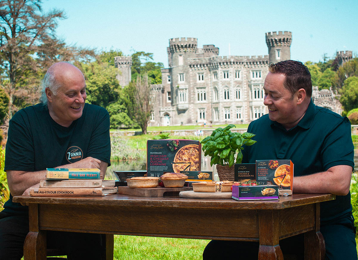 Owen Mullins and Neven Maguire sit at a wooden table in front of a majestic castle, showcasing various savoury pies, quiches and cookbooks.