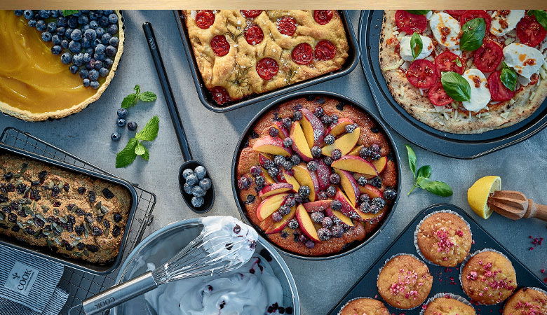 Overhead view of an artisanal baking spread featuring various homemade treats including a blueberry tart, rustic galettes with tomatoes, a fruit upside-down cake, pizza, muffins, and a mixing bowl, all arranged on a blue surface