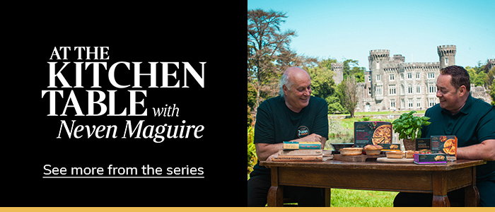 Neven Maguire sits at a table outdoors with Owen Mullins, surrounded by food products, with a historic castle visible in the background. At the Kitchen Table logo is to the right on a black background with a call to action of 'see more from the series'