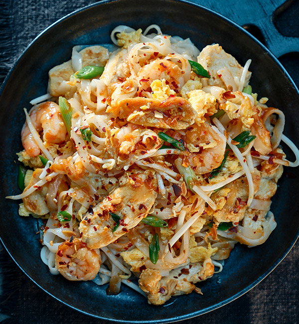 A close-up of a bowl of stir-fried noodles with shrimp, chicken, vegetables, and red pepper flakes, garnished with green onions.