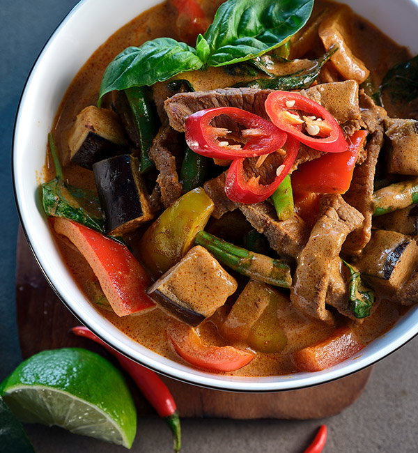 A bowl of Thai red curry with beef, eggplant, bell peppers, green beans, and fresh basil, garnished with red chilli slices.