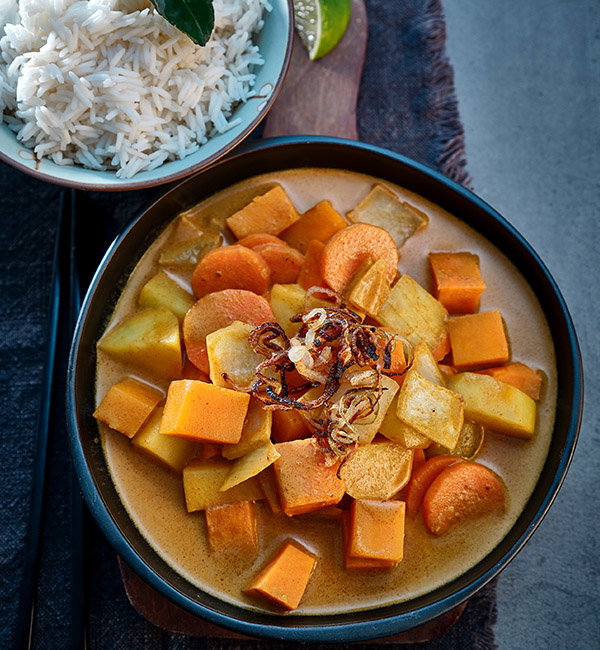 A bowl of colourful vegetable curry topped with fried onions, served alongside a small bowl of white rice.