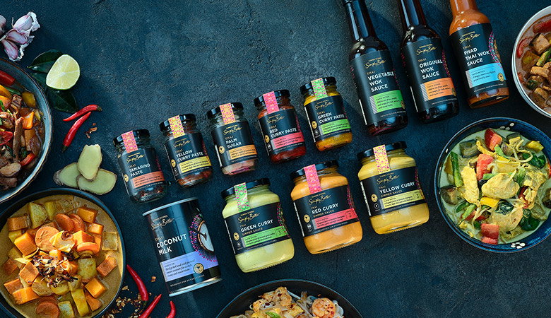 Selection of Simply Better Thai cooking ingredients and sauces arranged on a dark surface, including curry pastes, curry sauces, coconut milk and wok sauces, surrounded by prepared Thai dishes.