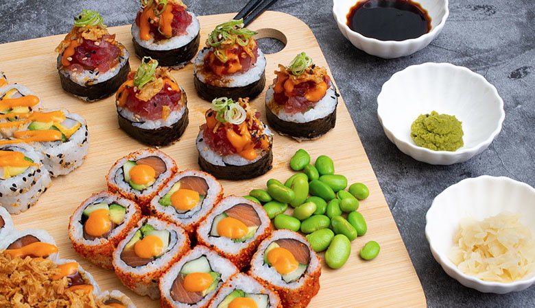 A wooden platter features an assortment of sushi rolls, edamame, and condiments like soy sauce, wasabi, and pickled ginger.