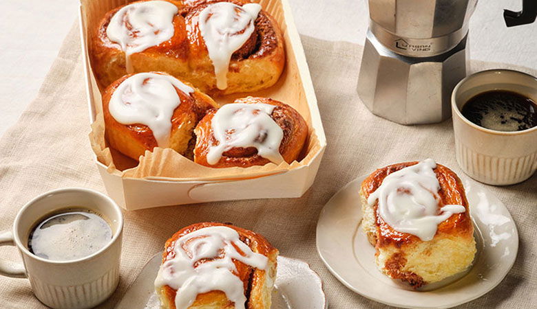 Freshly baked cinnamon rolls drizzled with icing, served with coffee in cups and a stovetop espresso maker, on a light tablecloth.